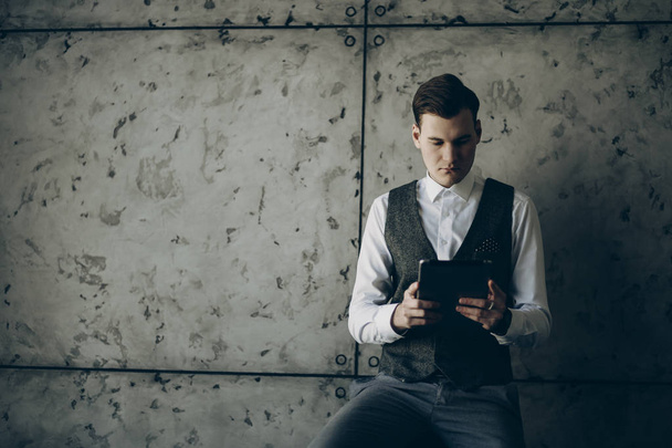 Retrato de jovem empresário sentado em uma cadeira operando em um tablet contra uma parede cinza no escritório
. - Foto, Imagem