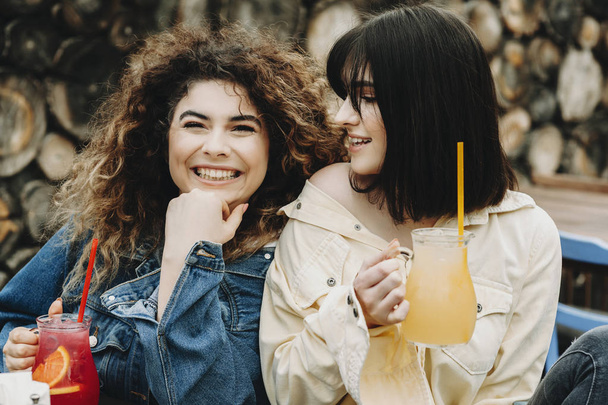 Porträt zweier kaukasischer Mädchen, die auf der Terrasse eines städtischen Restaurants sitzen und roten und gelben Saft in der Hand halten, während lockiges Mädchen vor dem Fotografen posiert und brünettes Mädchen sie lächelnd ansieht. - Foto, Bild