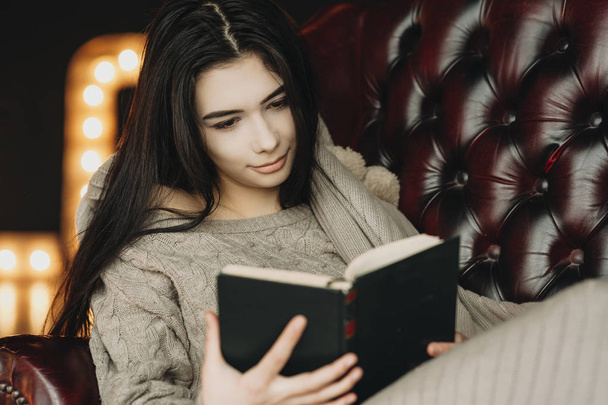 Roztomilá mladá bruneta sedí v koženém křesle a čtení knihy. - Fotografie, Obrázek