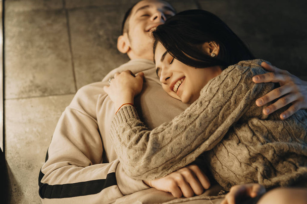 Πορτρέτο του ένα εκπληκτικό ζευγάρι που ακουμπά στο πάτωμα γελώντας ενώ κοπέλα κλίνει επάνω στο στήθος του ο φίλος με κλειστά μάτια κοντά σε θερμό φως. - Φωτογραφία, εικόνα