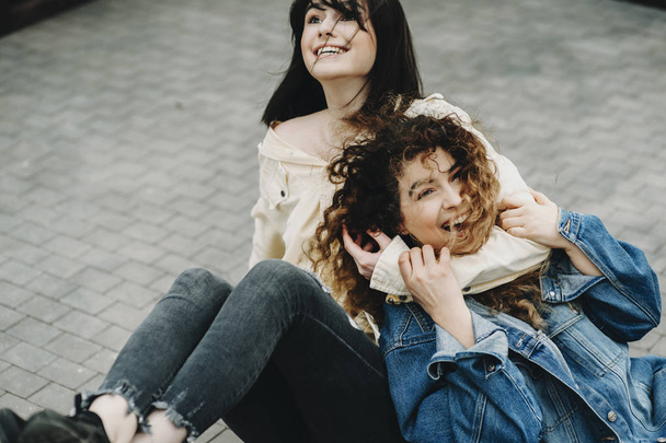 Charmante zwei Frauen amüsieren sich auf dem Boden einer Terrasse in der Stadt, wo Brünette den Hals ihrer Freundin hält und lacht, während lockiges Mädchen lachend in die Kamera blickt. - Foto, Bild