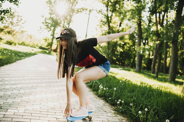 Ładna, młoda dziewczyna, jazda na deskorolce w parku podczas słuchania muzyki w słuchawkach. - Zdjęcie, obraz