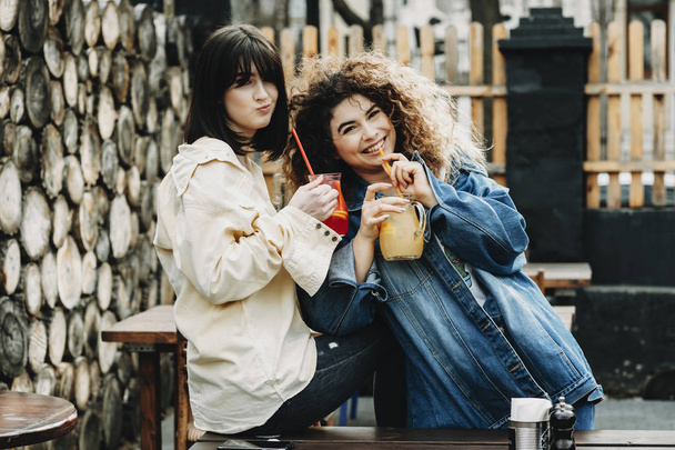 Two different caucasian girlfriends having fun in the city looking into camera and doing funny faces laughing and drinking ice juice. - Photo, image