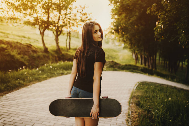Portrait from back of a amazing young girl posing at the camera and holding skateboard outdoor. - Fotografie, Obrázek