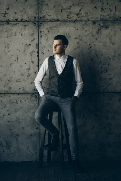 Full length portrait of a handsome young manager sitting on a chair with hands in his pocket and looking away thoughtfully against a grey wall. - Foto, Imagen