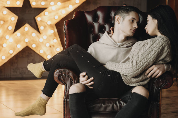 Giovane coppia caucasica seduta su una sedia di pelle e guardando sconvolto l'un l'altro mentre il ragazzo sta tenendo la sua ragazza sulle braccia, contro decorato con luci calde stella
. - Foto, immagini