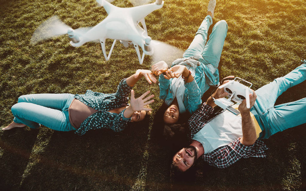 Grupa przyjaciół trzy różne rasy i płci: układanie na trawniku lato i nagrywanie wideo dla swoich blogów korzystających latający dron, który działa facet kaukaski za pomocą pilota zdalnego sterowania - Zdjęcie, obraz