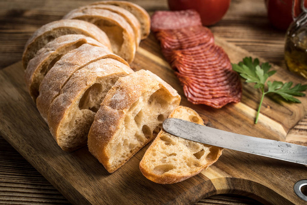 Italian ciabatta bread cut in slices on wooden chopping board with salami. - Photo, Image