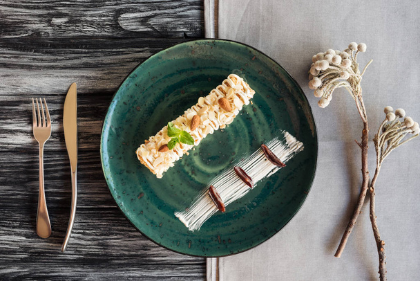 κάτοψη του γκουρμέ γλυκό επιδόρπιο με αμύγδαλα και δυόσμο σε πιάτο και πιρούνι με μαχαίρι σε ξύλινο τραπέζι  - Φωτογραφία, εικόνα