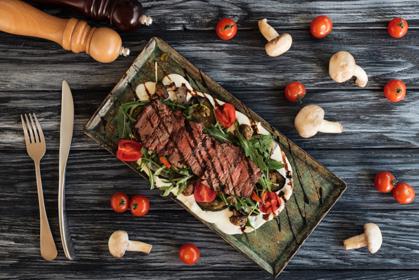 bovenaanzicht van heerlijke gekookte biefstuk met groenten, voorvork met mes en specerijen op de houten tafel   - Foto, afbeelding