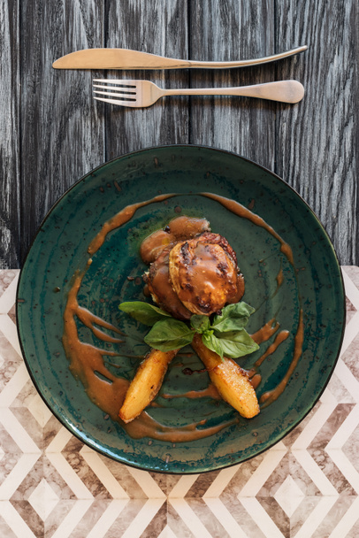 νόστιμο κρέας με τη σάλτσα και τα φύλλα βασιλικού, πηρούνι και μαχαίρι σε ξύλινο τραπέζι  - Φωτογραφία, εικόνα