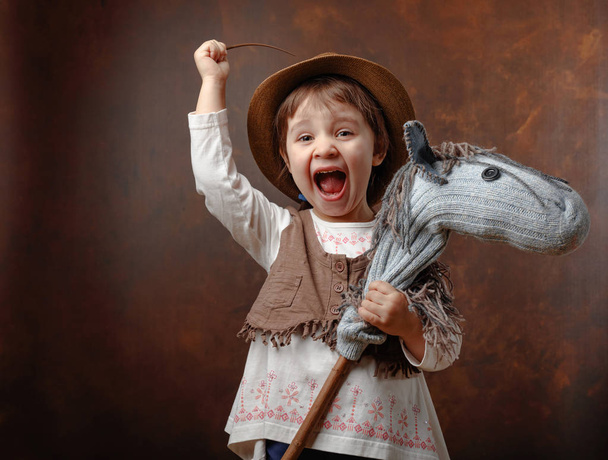Cute little girl dressed like a cowboy playing with a homemade horse. Expressive facial expressions. Copy space for your text. - Photo, Image