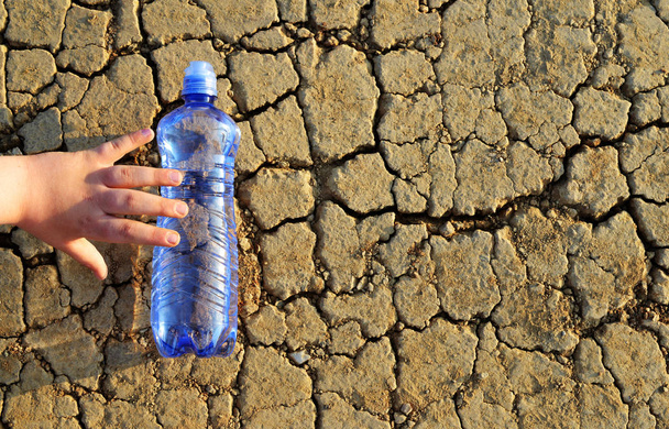 子供の手に達するひび割れ乾燥した土壌で横になっている水のボトル。地球温暖化の影響の概念. - 写真・画像