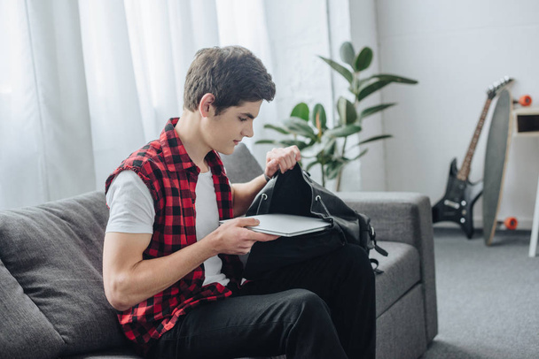 male teenager putting laptop into bag while sitting on sofa - 写真・画像