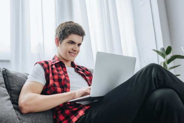 smiling teen boy typing on laptop while sitting on sofa  - Фото, зображення