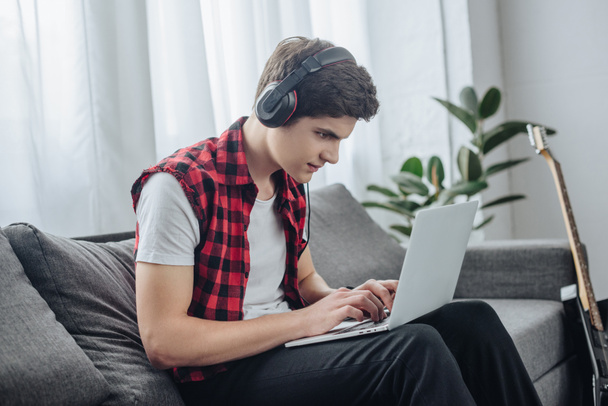 concentrated teenager with headphones playing game on laptop while sitting on sofa  - Photo, Image