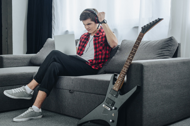 Teen chłopiec ze słuchawkami gra na laptopie siedząc na kanapie z gitara elektryczna w pobliżu - Zdjęcie, obraz