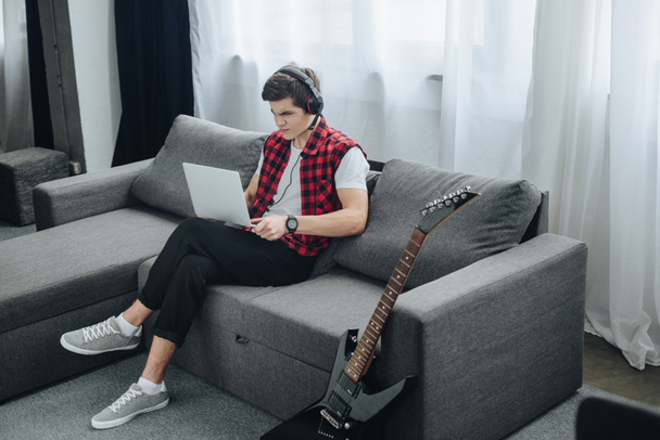 teenager with headphones playing game on laptop while sitting on sofa with electric guitar near - Photo, Image