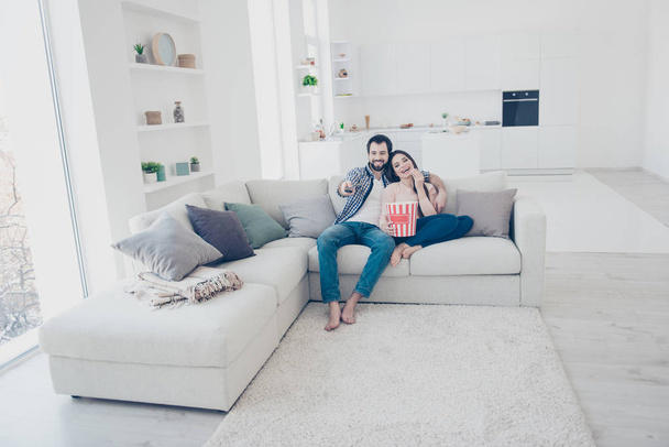 Full size portrait of stylish positive couple sitting in modern white apartment using console looking comedy funny channel program film on television hugging embracing eating snack - Fotografie, Obrázek