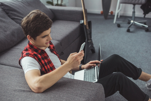 handsome teenager using smartphone and laptop while sitting on floor - Φωτογραφία, εικόνα