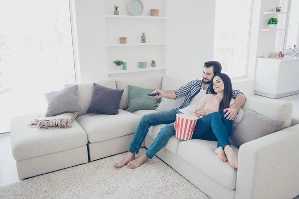 Portrait of trendy attractive couple sitting in modern living room using console trying to find interesting channel program film on television hugging embracing eating snack - Zdjęcie, obraz