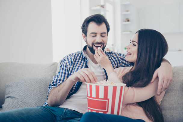 Portrait of cheerful positive couple sitting indoor in  livingroom having bucket of popcorn woman feeding boyfriend giving snack enjoying time together - Photo, image