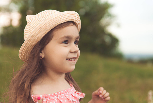 Beautiful little girl in straw hat with long hair looking away in summer field on sunset - Foto, Bild