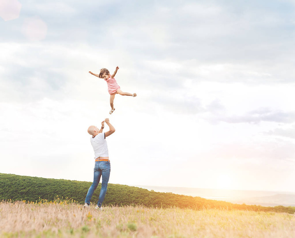 Happy Father playing with his daughter in sunny field on sunset. Summer happy family lifestyle, truly emotions. Little girl flying high in the sky from dad's hands - 写真・画像