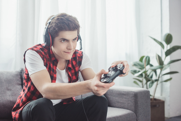 έφηβο με ακουστικά που παίζει video game με gamepad στο σπίτι - Φωτογραφία, εικόνα