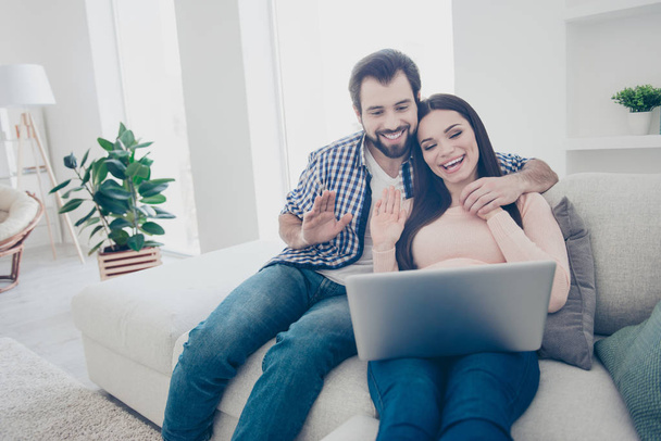 Portrait of friendly couple having using electronic devices gadget internet wifi for social networks  having video call with relatives parents friends, hugging embracing sitting indoor - Zdjęcie, obraz