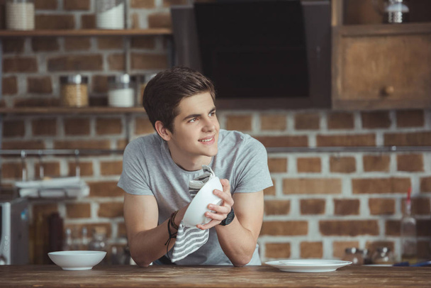 smiling teen boy cleaning dinnerware with towel on kitchen - Zdjęcie, obraz