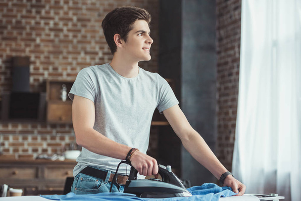 dreamy caucasian teenager ironing blue shirt at home - 写真・画像