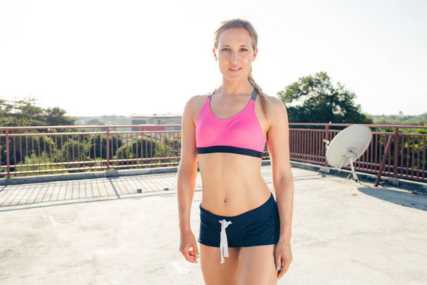 smiling young young sportswoman standing and looking at camera on rooftop - Photo, Image