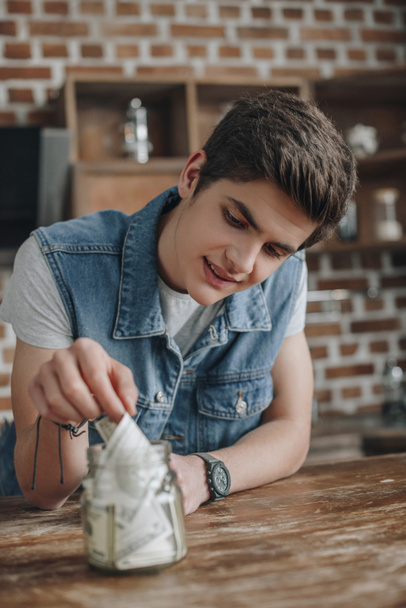 handsome teenager putting dollar banknotes into saving glass jar for money - Photo, Image