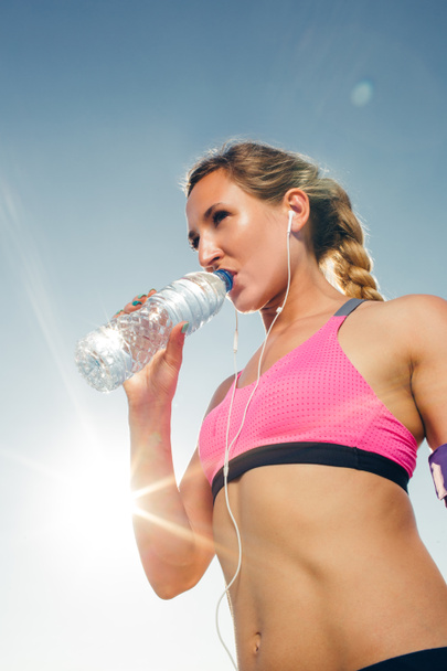 Tiefansicht einer jungen Sportlerin mit Kopfhörern, die vor blauem Himmel Wasser aus der Flasche trinkt - Foto, Bild
