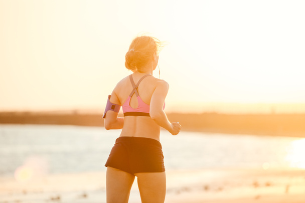 日光に対してビーチでジョギング ランニングのアームバンド ケースにスマート フォンをイヤホンでスポーツウーマンの背面図  - 写真・画像