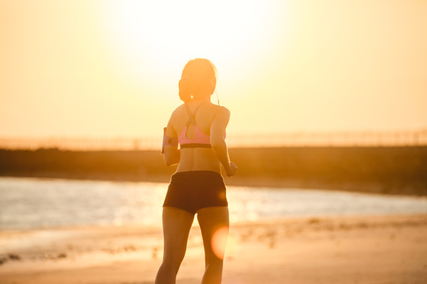rear view of silhouette of sportswoman in earphones with smartphone in running armband case jogging on beach against sunlight  - Foto, Bild