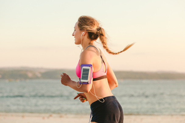 vue latérale de la sportive souriante dans des écouteurs avec smartphone dans le cas de course brassard jogging sur la plage avec mer derrière
  - Photo, image