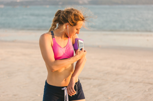 молода спортсменка в навушниках, використовуючи смартфон у футлярі для пов'язки на пляжі з морем позаду
  - Фото, зображення