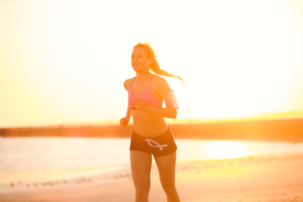 silhouette of sportswoman in earphones with smartphone in running armband case jogging on beach against sunlight - Photo, Image