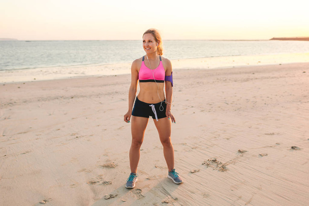 усміхнена спортсменка в навушниках зі смартфоном у футлярі, що стоїть на піщаному пляжі з морем позаду
  - Фото, зображення