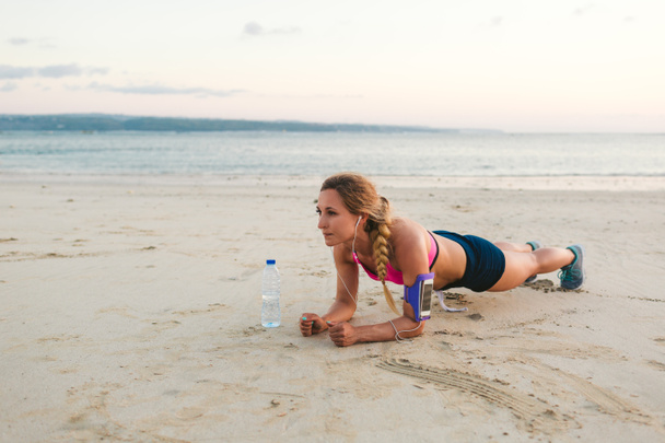 sportswoman in earphones with smartphone in armband case and bottle of water doing plank on sandy beach  - Фото, изображение