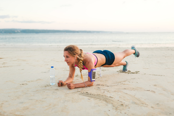 young sportswoman in earphones with smartphone in running armband case and bottle of water doing plank on sandy beach  - Photo, Image