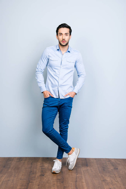 Full size fullbody portrait of harsh cool bachelor with black hair holding two hands in pockets of jeans looking at camera standing over grey background - 写真・画像