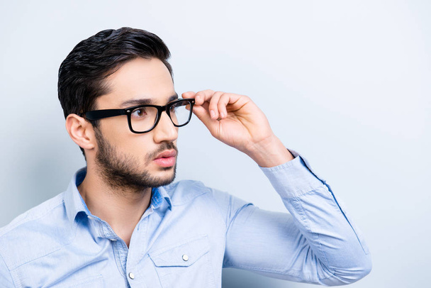 Portrait of virile trendy bachelor with black hair in blue shirt casual outfit looking away holding eyelet of glasses on face isolated on grey background - Photo, image