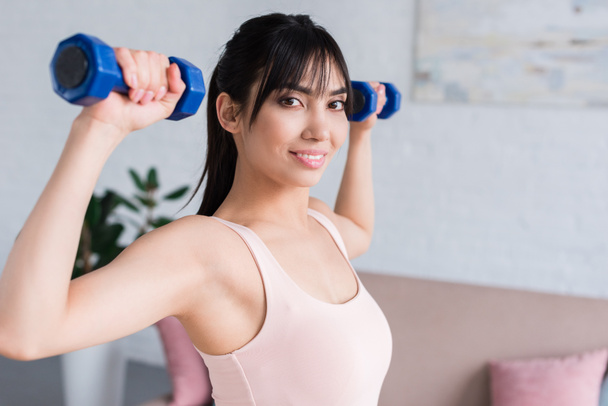 happy young woman working out with dumbbells at home and looking at camera - Fotografie, Obrázek