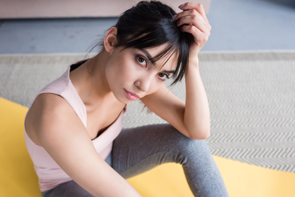 high angle view of beautiful young woman sitting on yoga mat and looking at camera at home - Photo, Image