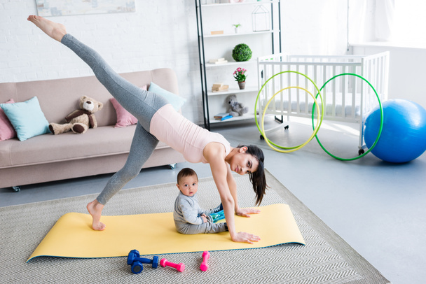 young mother practicing yoga in One Legged Downward-Facing Dog pose while her little child sitting on mat at home - Foto, Imagen