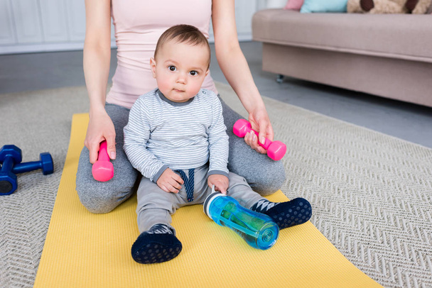 cropped shot of mother with dumbbells and little child sitting on yoga mat at home - Photo, Image