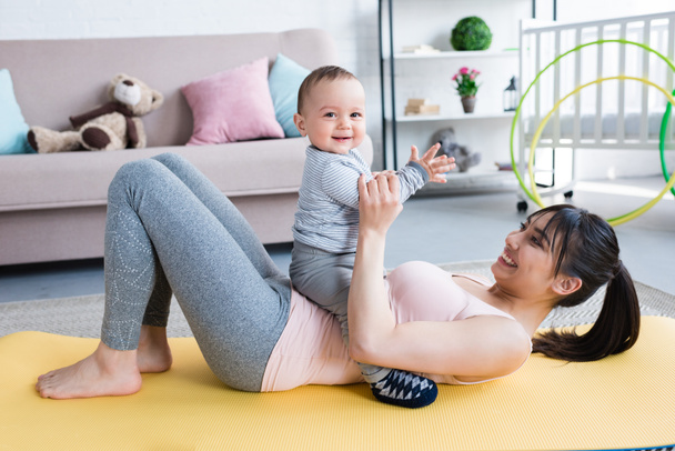 young beautiful mother lying on yoga mat with her little child at home - Photo, image